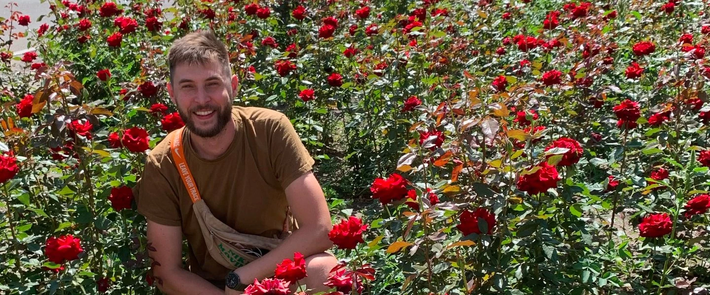 Ein hellhäutiger Mann mit Bart kniet lächelnd inmitten eines üppigen Beets voller roter Rosen. Er trägt ein braunes T-Shirt u