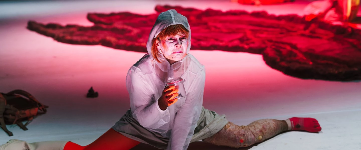 Eine hellhäutige Frau sitzt im Spagat auf einer Theaterbühne und hält ein Glas. Sie trägt helle Kleidung, rote Strümpfe und a