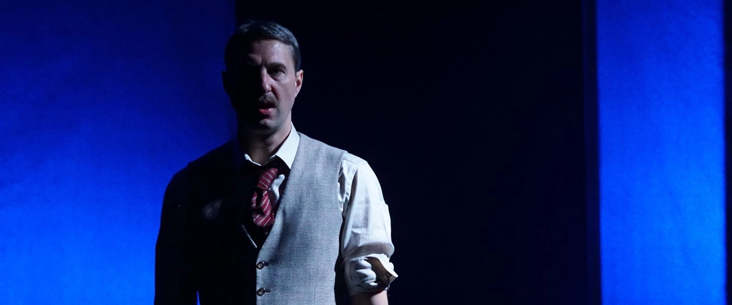 Ein Schauspieler steht halb im Dunkeln auf der Bühne, nur von einem kalten blauen Licht seitlich beleuchtet. Er trägt einen g